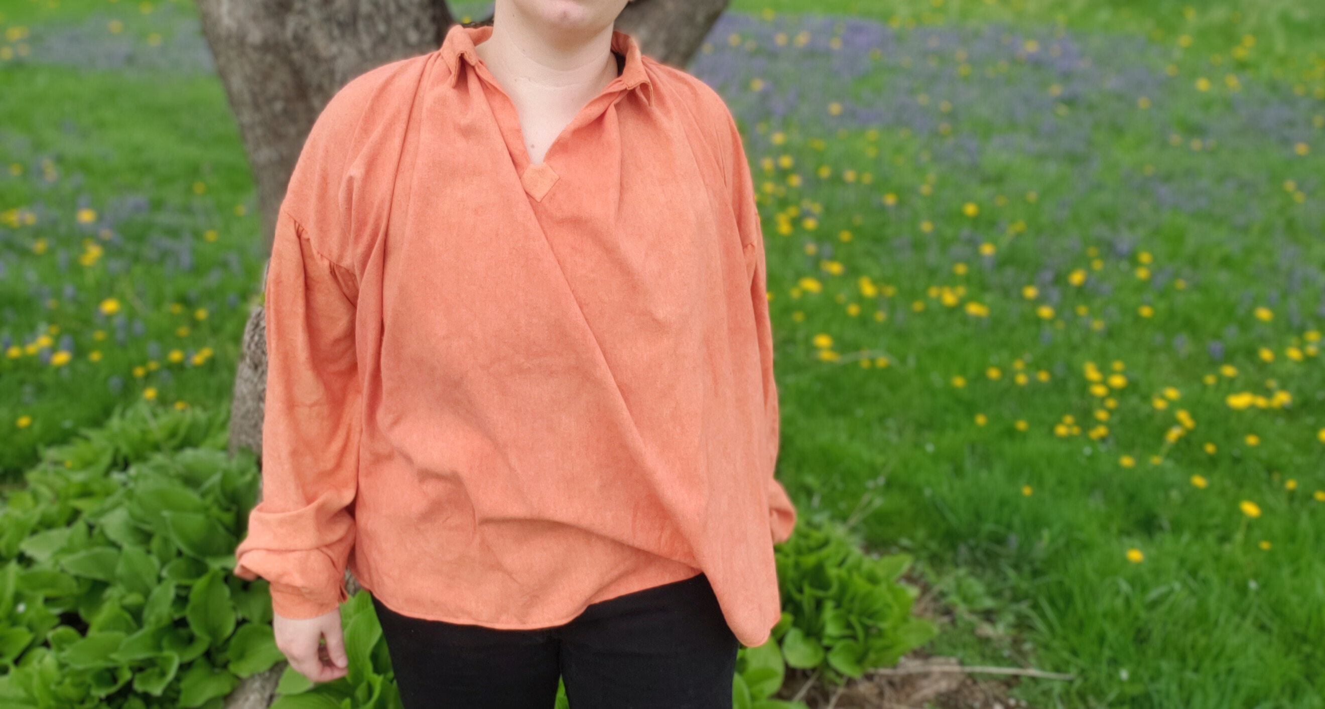 A white figure standing in a field of green grass, yellow, dandelions and purple flowers near a tree. The figure is wearing an orange 18th century shirt with poofy sleeves.