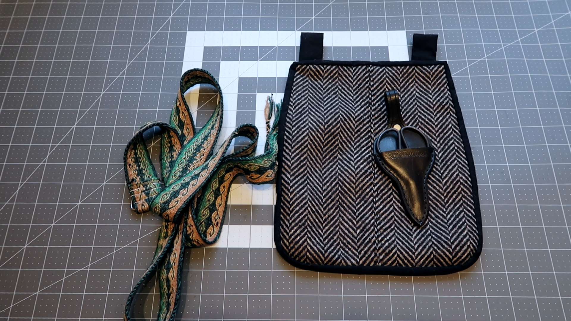 A grey cuttin mat background with a tablet woven belt and wool 18th century pocket. The belt is woven in a ram's head pattern in dark green, light green, yellow, and pink wool. The pocket is made with a tan and black herringbone pattern with black binding. Atop the pocket is a leather holster with a pair of small scissors.